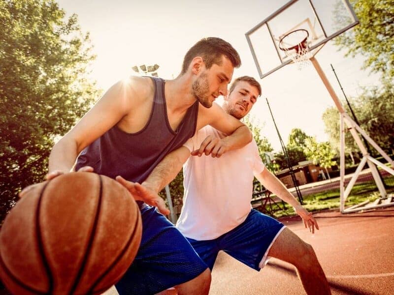 Man in maroon tank top elbows man in white t-shirt during basketball at the park.