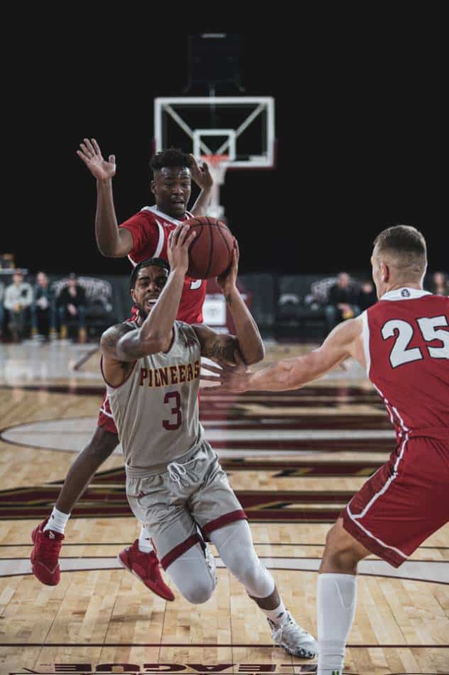 College basketball player in silver and red drives to the basket against players in red.