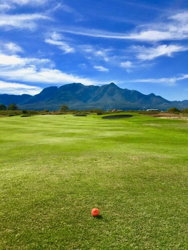 An orange golf ball sits on the fairway with mountains in the background.