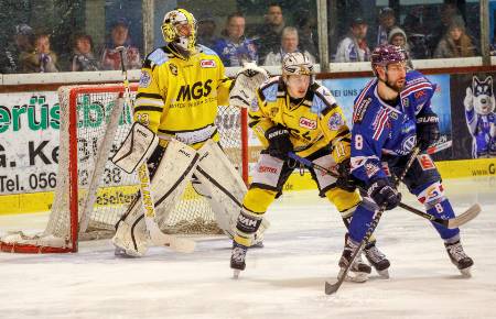 The goalie looks on as two opposing players jostle for position in front of the net.