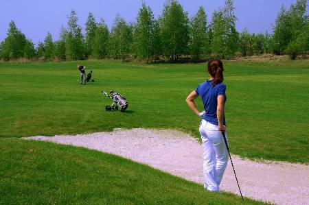 A female golfer looks on as another golfer takes a shot from the fairway.